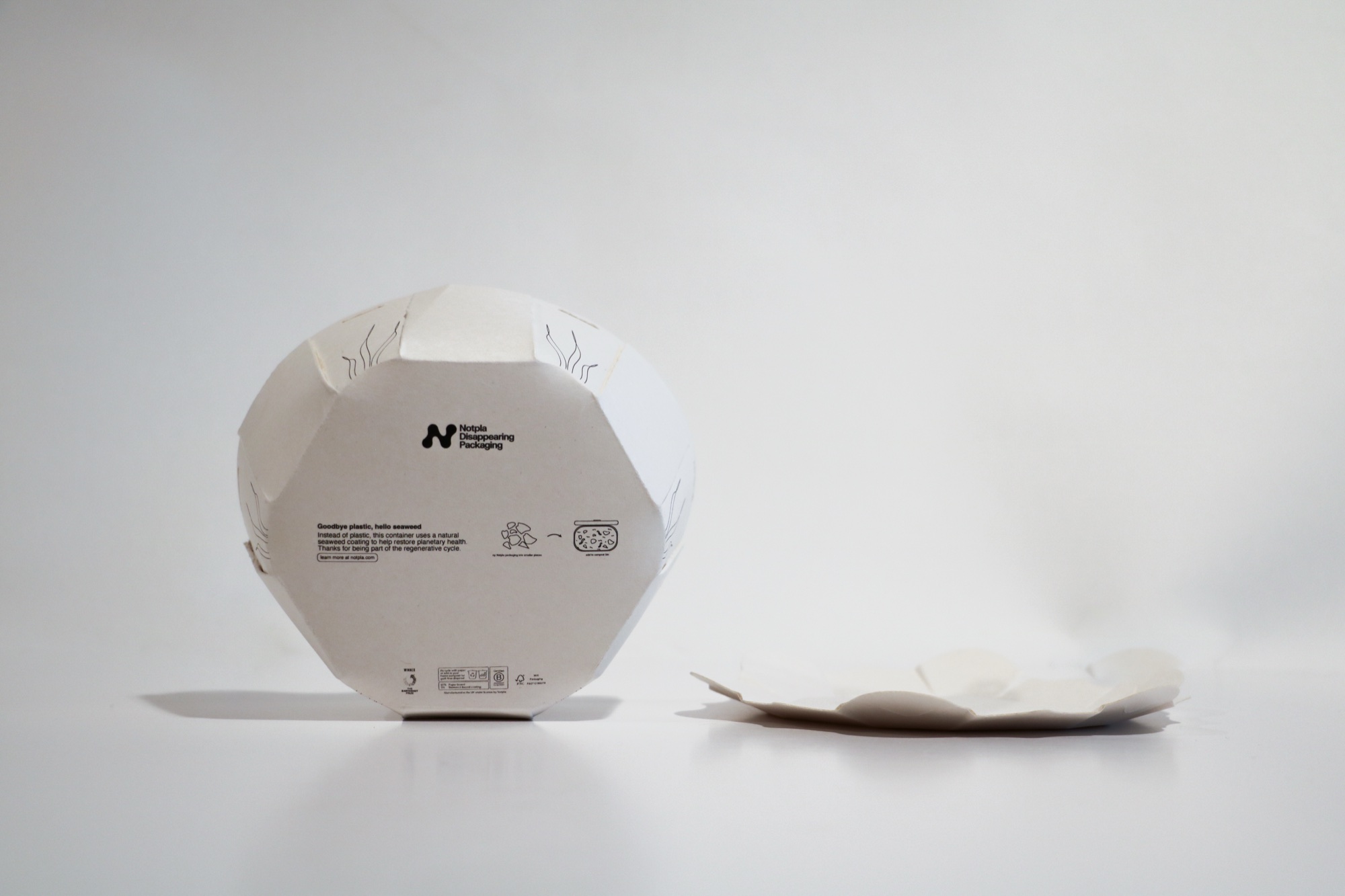 A closed Nelumbo container standing on its side, showing the printed Notpla base, beside a lid